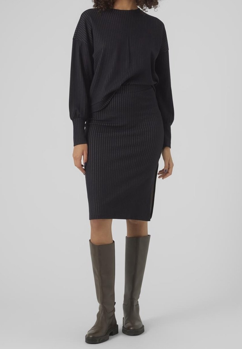 Femme portant un top noir à fines rayures manches longues et une jupe assortie fendue sur le côté, associée à des bottes montantes en cuir marron.