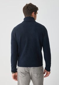 Pletený tmavomodrý sveter s vysokým rebrovaným golierom. Má textúrovanú tkaninu a rebrované manžety, skombinovaný so sivými džínsy.