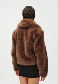 Καφέ ψεύτικο σακάκι γούνας με φαρδύ γιακά και μακριά μανίκια. Μαλακή υφή και όγκος στο σχέδιο. Φοριέται με μια μαύρη εφαρμοστή φούστα.