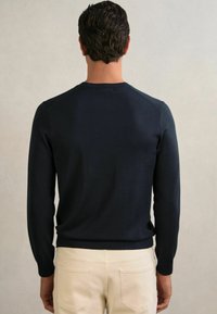 Maglione a maglia blu navy con maniche lunghe e polsini a coste. La vista posteriore mostra una forma aderente e un design delle spalle senza cuciture, abbinato a pantaloni chiari.
