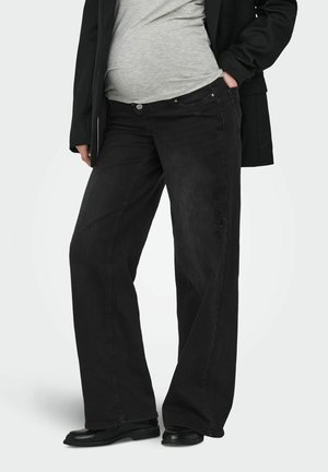 Osoba oblečená v čiernych džínsach s širokými nohavicami a jemným opotrebením, šedej košeli, čiernej bunde a čiernych topánkach, stojaca s jednou rukou vo vrecku.