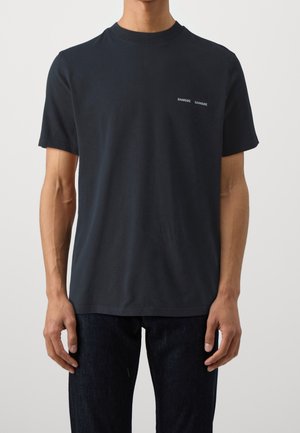 Mann trägt ein schlichtes schwarzes T-Shirt mit kurzen Ärmeln und kleinem weißem "SAMSOE SAMSOE"-Schriftzug auf der linken Brust, kombiniert mit dunkler Hose.