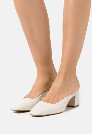 Voeten in witte instap block heel schoenen met een spitse neus op een effen witte achtergrond.