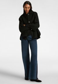 Chaqueta de lana negra de corte oversize con cuello y botones plateados, combinada con jeans de denim azul de pierna ancha con aberturas laterales.