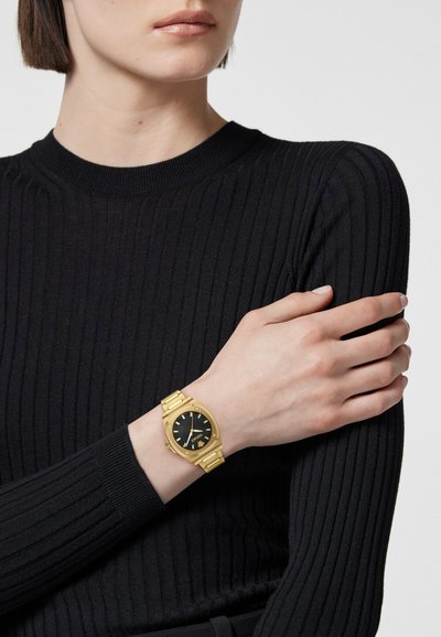 Personne portant un pull côtelé noir, les bras croisés, montrant une montre bracelet en or avec un cadran noir au poignet gauche.