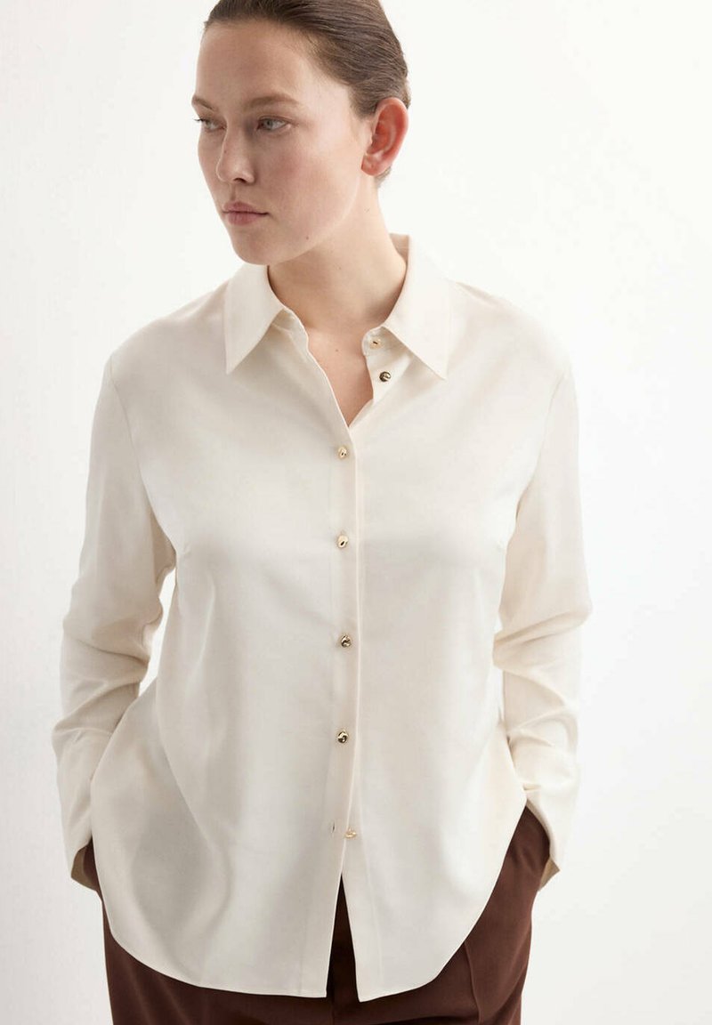 Femme portant une chemise crème à manches longues boutonnée, les mains dans les poches d’un pantalon marron, regardant sur le côté sur un fond blanc.