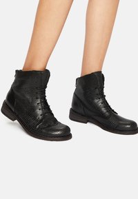 Botas de tobillo de cuero negro con un diseño perforado, cordones y un panel lateral texturizado; terminadas con un tacón de madera bajo.
