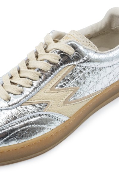 Scarpa da ginnastica bassa con tomaia testurizzata in argento metallico, dettaglio fulmine beige, lacci color crema e suola traslucida color gomma.