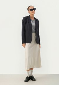 Väärennetty tumman sininen pinstripe-takki harmaan t-paidan päällä, beige midi-hame, valkoiset sukat ja mustat loafersit. Suuret mustat aurinkolasit.