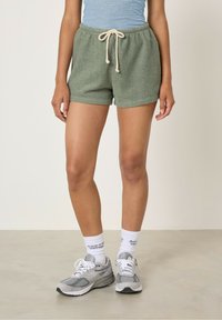 Shorts in terry verde con vita elastica e cordino beige, caratterizzati da una vestibilità casual e tasche laterali. Abbinati a sneakers grigie.