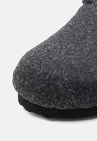 Nærbillede af en mørkegrå filt hjemmesko med tekstureret stofoverdel og sort gummisål på en hvid baggrund.