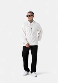 Weiße Zip-up-Jacke mit hohem Kragen, schwarze Hose und weiße Sneakers. Die Accessoires umfassen dunkle Sonnenbrillen. Minimalistisches Design mit weicher Textur.