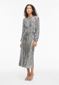 Silverglittrig klänning med långa ärmar och rak silhuett. Har en rundad halsringning och subtilt skimmer, kombinerad med matchande silverfärgade sandaler.