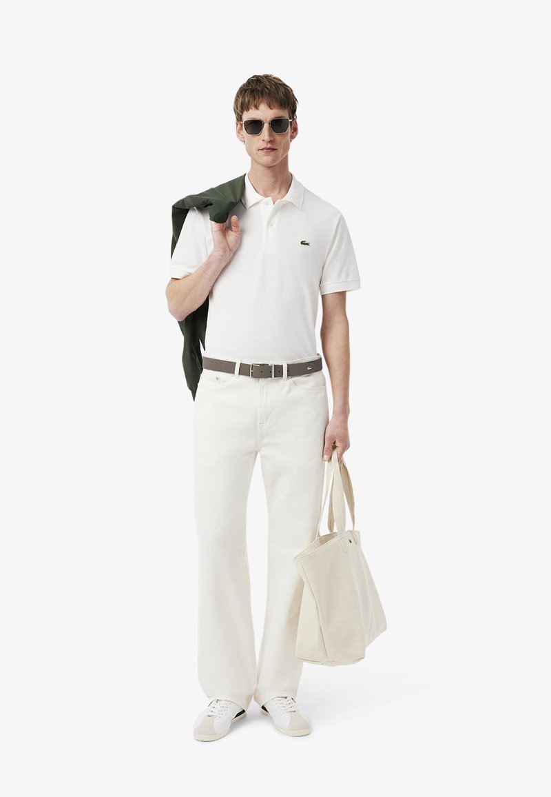 Hombre vistiendo polo blanco y pantalones, gafas de sol, sosteniendo una bolsa de lona beige en una mano y una chaqueta verde sobre el hombro con la otra.