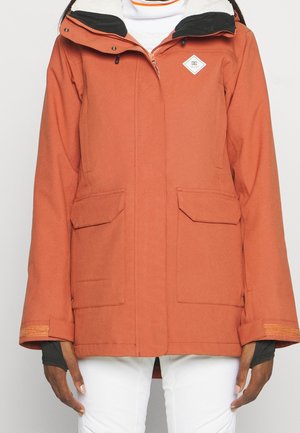 Kurtka w kolorze rdzawego pomarańczu z przednim zamkiem błyskawicznym, dwie kieszenie z patką, czarna wewnętrzna podszewka kaptura i białe spodnie, noszona przez osobę o ciemnej karnacji.