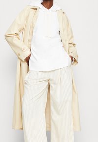 Un trench coat crema sopra una felpa bianca, abbinato a pantaloni a righe sottile color avorio. Il tessuto è morbido; presenta una vestibilità rilassata e dettagli con bottoni.
