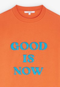 Felpa arancione con scollatura a girocollo. Presenta un testo ricamato blu "GOOD IS NOW" ben visibile sulla parte anteriore.