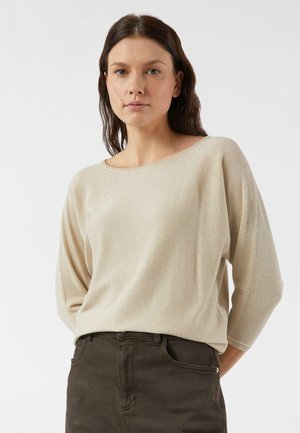 Beige, funkelnder Strickpullover mit weitem Rundhalsausschnitt und dreiviertellangen Ärmeln, kombiniert mit dunkelbrauner Hose. Weiche Textur mit lockerem Schnitt.