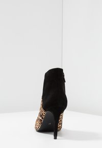 Bota de tacón alto de ante negro con estampado de leopardo, vista desde atrás sobre una superficie blanca y un fondo blanco liso.
