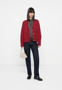 Rød kabelstrikket cardigan med gullknapper, båret over en svart polkadotbluse og kombinert med mørke jeans og svarte loafers.