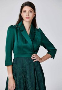 Femme aux longs cheveux bruns portant un haut en satin vert foncé de style blazer et une jupe florale texturée, posant avec la main sur la taille contre un fond clair.