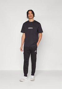 Juoda medvilninė marškinėliai su baltu „adidas“ logotipu, derinami su juodomis kargo kelnėmis su šoninėmis kišenėmis ir baltais logotipo akcentais, dėvimi su pilkais sportbačiais.