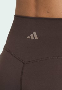 Hnedé športové legíny s vysokým pásom, s hladkou textúrou a vystupujúcim sivým logom Adidas blízko pása.