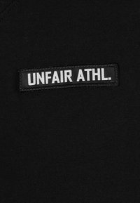 Camiseta de algodón negra con un parche rectangular de cuero que lleva el texto "UNFAIR ATHL." en blanco. Diseño sencillo de cuello redondo, textura suave.