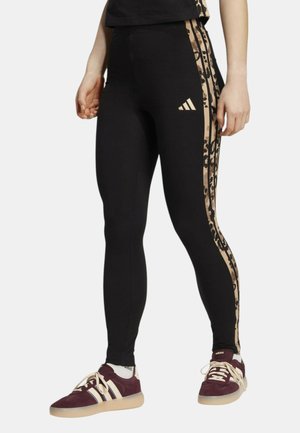 Modella che indossa leggings neri con bande laterali beige con stampa animalier e logo Adidas, abbinati a sneakers Adidas color bordeaux e calzini bianchi.