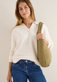 Cremfarbener V-Ausschnitt-Strickpullover mit gerippten Bündchen, kombiniert mit dunkelblauen Jeans. Hält eine olivgrüne, gepolsterte Schultertasche.