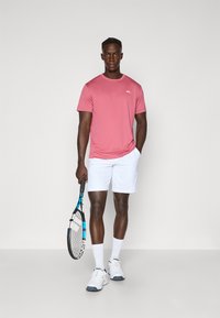 Rozā sporta krekls ar diskrētu logotipu, baltas šortus, baltas tenisa kurpes un rokās turam tenisa nūju ar zilām akcentiem.