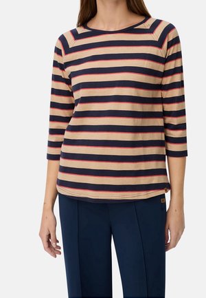 Femme portant une chemise beige avec des rayures horizontales bleu marine et rouge, et un pantalon bleu marine, debout devant un fond uni.