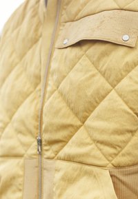 Prešívaná žltá bunda s celým zipsom, textúrovanou látkou a suédovou nášivkou so zapínaním na dve patentky na hrudi.