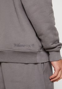 Szary sweatshirt z prążkowanymi mankietami i dołem, z haftowanym napisem "Milano 1978" w pobliżu dolnej krawędzi. Miękka, strukturalna tkanina.