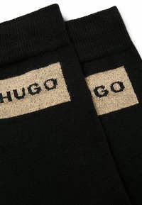 Calzini neri con polsino a coste, che presentano una toppa rettangolare dorata con la scritta 'HUGO' tessuta in nero. Texture liscia, design moderno.