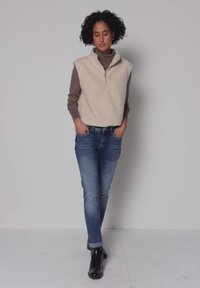 Femme aux cheveux bouclés portant une veste en polaire beige sur un pull à col roulé marron, un jean bleu et des bottines noires, debout contre un mur clair uni.