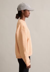 Brzoskwiniowa bluza z luźnym krojem i ściągaczami, połączona z czapką w czarno-białą kratę. Model stoi w profilu na tle neutralnym.