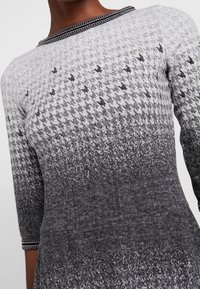 Haut gris tricoté avec un motif pied-de-poule, effet dégradé du clair au foncé et bordure décorative noire le long de l'encolure.