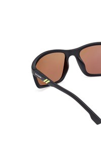 Gafas de sol de plástico negro con lentes marrones, con un diseño geométrico. Las patillas tienen rayas de acento amarillas y el logotipo de la marca.