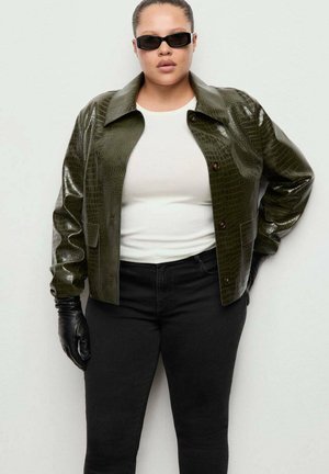 Persona che indossa una giacca di pelle verde scuro con texture, camicia bianca, pantaloni neri, guanti neri e occhiali da sole neri stretti, in piedi davanti a uno sfondo uniforme.