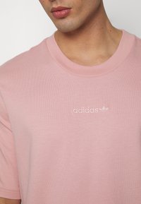 Vaaleanpunainen puuvillapaita, jossa on pyöreä kaula-aukko, lyhyet hihat ja kirjailtu "adidas" -logo rinnassa; sileä rakenne, rento malli.