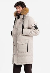 Parka beige imbottita con cappuccio foderato in pelliccia, tasche frontali e dettagli neri sulle maniche. Presenta una chiusura con zip e cordini regolabili.