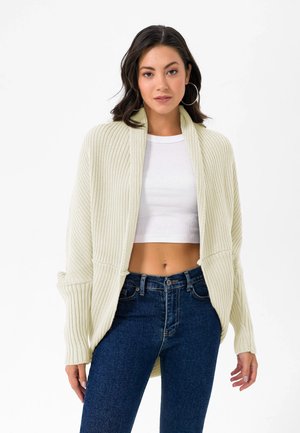 Cremefarvet strik cardigan med ribbet tekstur, åbent frontdesign og lange ærmer, parret med en hvid crop top og mørkeblå jeans.