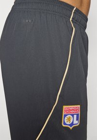 Mørkegrå atletikshorts med elastisk talje, prydet med en guldstribe og et broderet Olympique Lyonnais-logo.