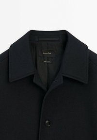 Manteau en laine noire avec un col pointu et un seul bouton visible, marqué "Massimo Dutti", fabriqué en Égypte.