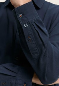 Navyblauwe langermige shirt met knoopmanchetten, textuurstof en contrasterende oranje knoopaccenten op de mouwen.