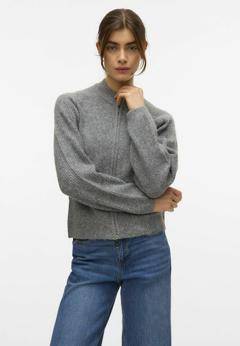 Suéter gris de punto con cuello alto y mangas acanaladas, con cierre de cremallera y corte relajado, combinado con jeans azules.