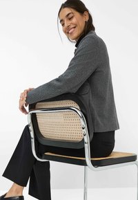 Donna sorridente con capelli scuri, in un blazer grigio e pantaloni neri, si siede di lato su una sedia in canna e cromata contro uno sfondo bianco.
