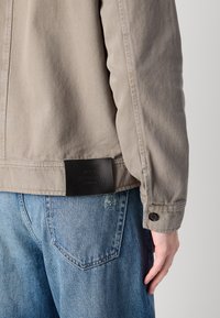Nærbilde av en person som har på seg en lys beige jakke og blå jeans, med en svart etikett på jakken nederst på ryggen og hånden hvilende ved siden av.