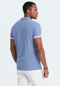 Světle modré polo tričko s klasickým límečkem, bílým lemem na rukávech a nenápadným logem poblíž dolního lemu; hladká bavlněná látka a přiléhavý střih.